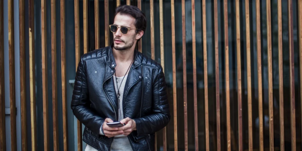 Leather and Fur Cleaning: Man wearing a stylish black leather jacket, holding a phone, and leaning against a wall with vertical wooden slats.