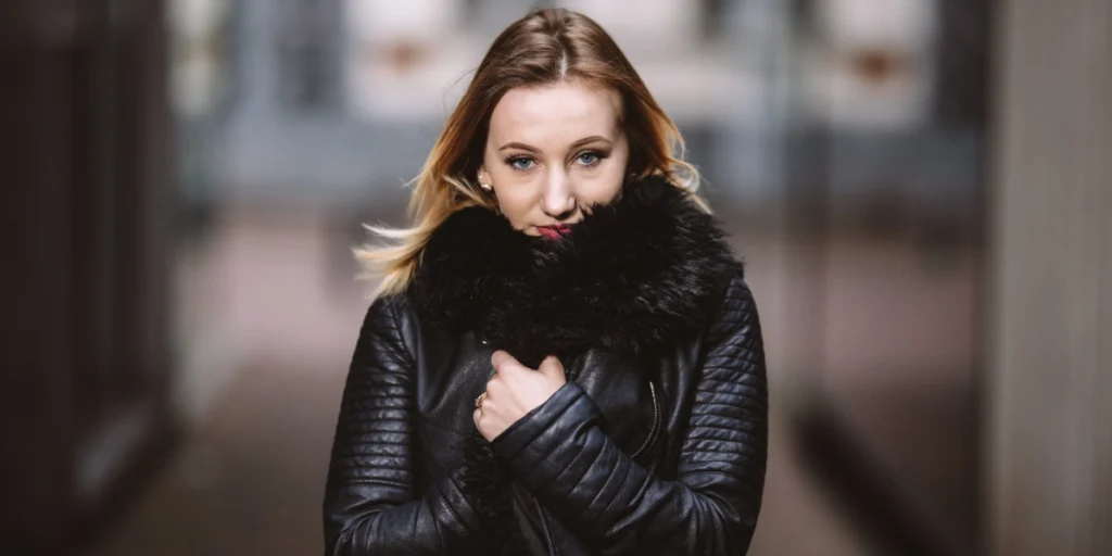 Leather and Fur Cleaning: Woman in a black leather jacket with a fur collar, holding it close to stay warm in an urban setting.